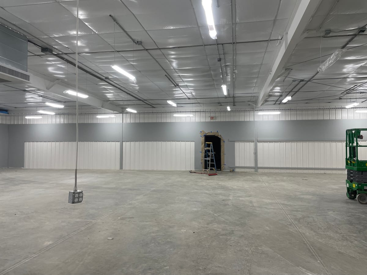 Steel-framed retail building interior mid-construction with a ladder, a scissor lift, and a taped-off containment area along the back wall