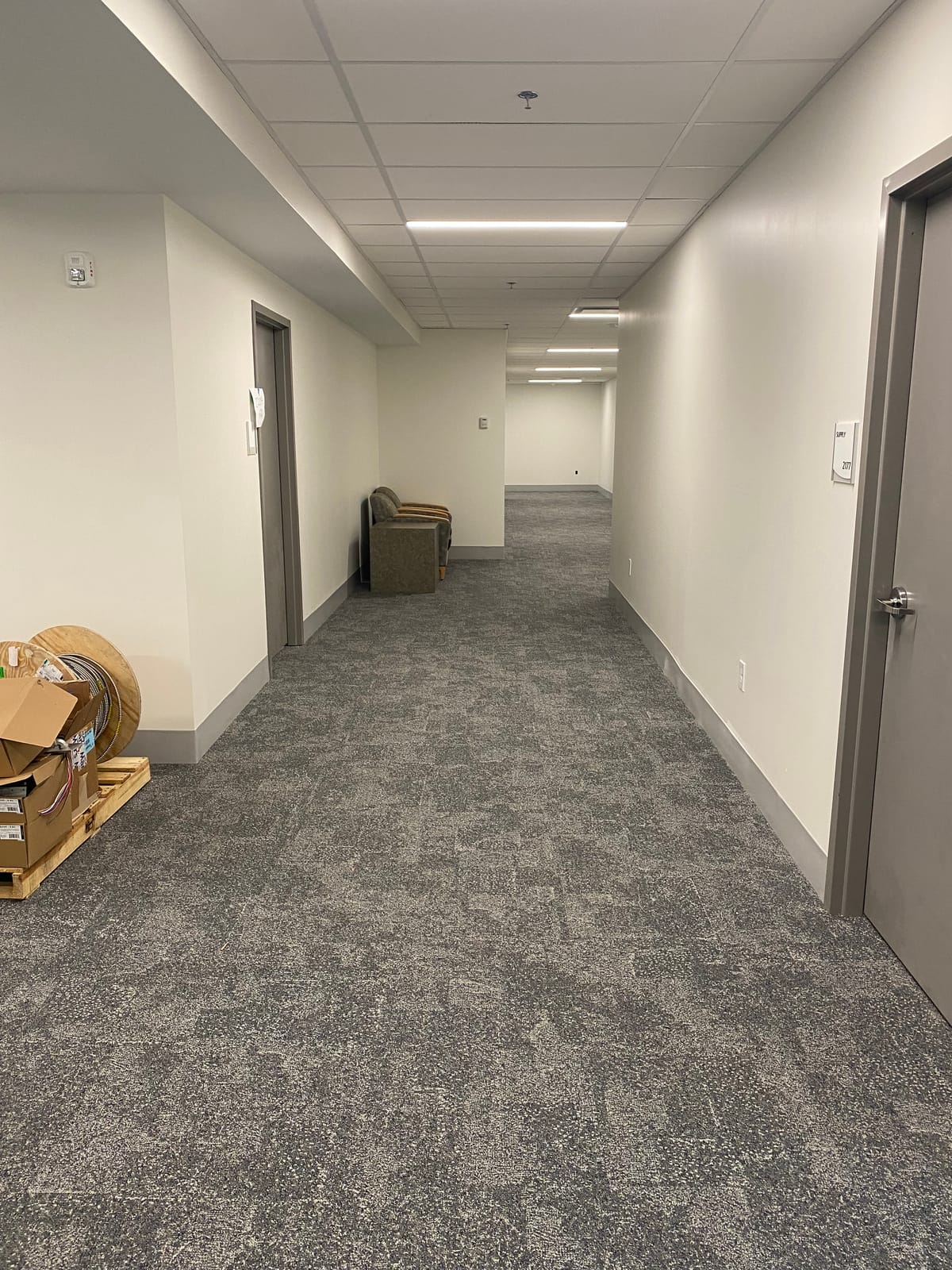 Finished office corridor with a 2x2 suspended acoustical ceiling grid and tile, recessed lighting, a sprinkler head, and a labeled supply closet door