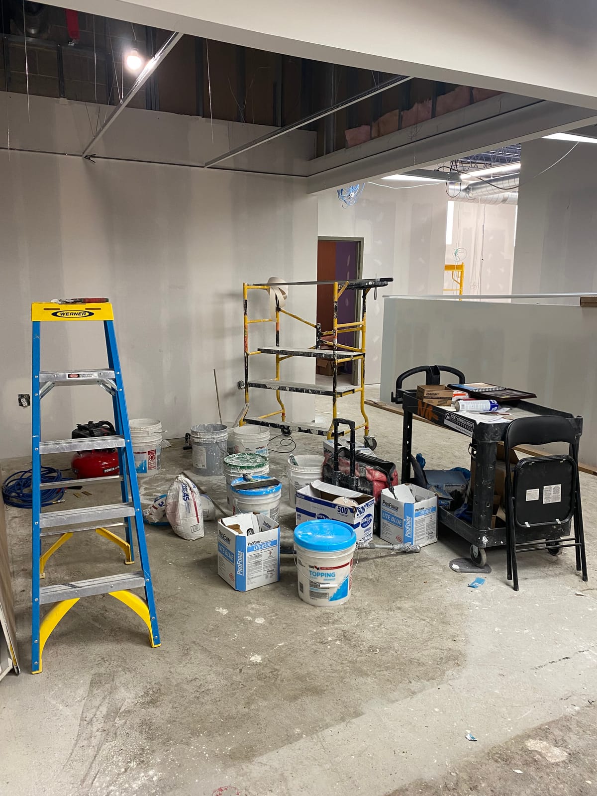 Rolling scaffold and buckets of joint compound and topping compound staged beneath a taped drywall ceiling soffit for finishing and skim-coat work