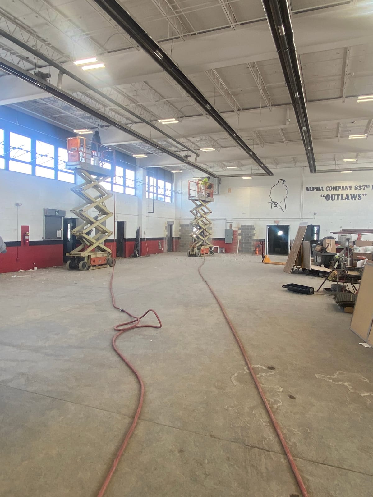Interior drill hall at a National Guard armory mid-repaint, scissor lifts at the wall below the Alpha Company 837th BEB mural